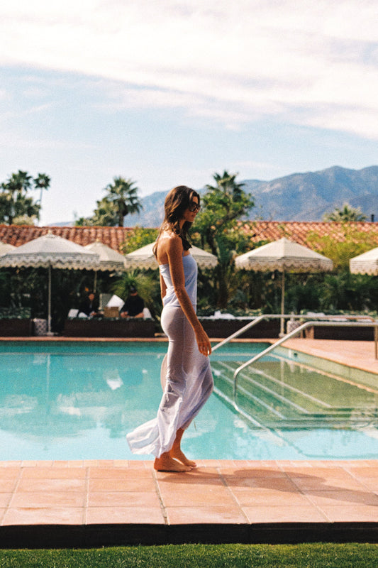 A woman in a light blue mesh cover-up dress walks barefoot beside a swimming pool at a resort, with umbrellas, palm trees, and mountains in the background.