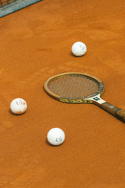 A vintage tennis racket with a brown handle lies on a clay tennis court next to three white tennis balls, with the court net visible in the background.